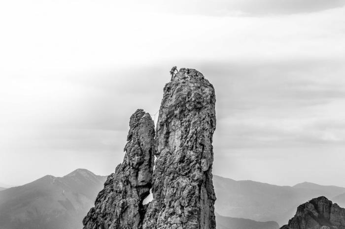 rojaus kraštovaizdis, svajonių peizažas, uolos pilkos spalvos, alpinistas užlipa ant aukščiausios uolos vaizde, nespalvota nuotrauka, pilkų kalnų siluetai horizonte